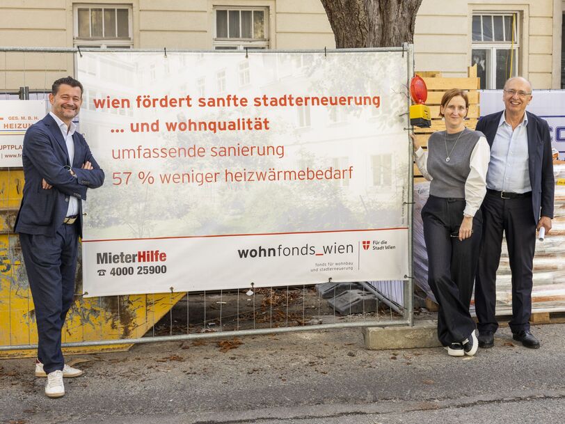 Drei Personen stehen vor einem Bauzaun mit einem Banner, das Informationen zur Stadtentwicklung und Wohnqualität in Wien zeigt